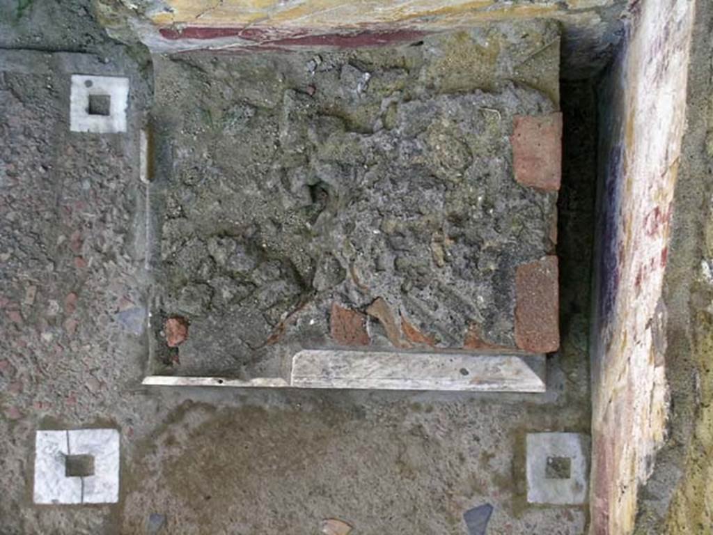 V.7 Herculaneum. May 2005. Looking down onto lararium in north-west corner of atrium. Photo courtesy of Nicolas Monteix.
According to Maiuri –
“E accanto alla cella ostiaria nell’angolo dell’atrio, come piu frequentemente a Pompei, troviamo invece per la prima volta a Ercolano il misero avanzo del podio di un Larario. Costruito e addossato posteriormente alla decorazione murale, forse dall’ultimo proprietario dell’abitazione, sarebbe stato, se fosse stato risparmiato dalla spoliazione e demolizione dei cavamonti dei cunicoli, il piu singlare e piu prezioso Larario che si potesse avere di Pompei e di Ercolano. Poiche non solo era rivestito di marmi, come il singolarissimo Larario di Lucio Cecilio Giocondo a Pompei, ma dal ricupero che si era fatto, fra le sue malconce rovine, di due monocromi marmorei, uno dei quali reca la firma di quello stesso artista Athenaios che appare sull’altro famoso monocromo ercolanese delle Giuocatrici di astragali, si era indotti a supporre che il Larario fosse ornato di pinakes marmorei dipinti nella preziosa tecnica dei monocromi ercolanesi. (Vol II, le Opere d’arte).  
(And next to the “cella ostiaria” (porter’s room) in the corner of the atrium, as more frequently in Pompeii, we find for the first time in Herculaneum the miserable remains of the podium of the Lararium. Built and leaning against the wall decoration, perhaps by the last owner of the house, it would have been, if it had been spared from the spoliation and demolition of the searchers in the Bourbon tunnels, the most singular and most precious Lararium that could be had in both Pompeii and Herculaneum. Since it was not only covered with marble, like the very singular Lararium of Lucius Caecilius Giocondo in Pompeii, but from the recovery that had been made, among its battered ruins, of two marble monochromes, one of which bears the signature of the same artist “Athenaios”, which appears on the other famous Herculaneum monochrome of the Astragali Players, it was led to suppose that the Lararium was adorned with marble “pinakes” painted in the precious technique of Herculaneum monochromes. (Vol. II, The Works of Art)
See Maiuri A., 1958. Ercolano I Nuovi Scavi (1927-1958) Vol. 1. Roma: Istituto Poligrafico dello Stato, (p.394-5).

