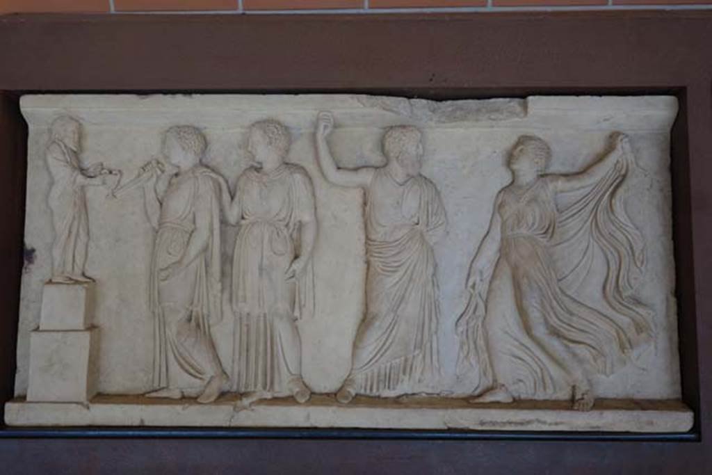 House of Dionysiac Reliefs, Herculaneum. June 2014. Second marble relief. Inventory number 88091.
Photo courtesy of Michael Binns.
This shows a dancing Maenad and a bearded man, probably Dionysius, facing each other on the right side.
On the left two female figures stand before an archaic Greek sculpture of Dionysius who is holding a kantharos. 
The adult woman has her hand on the shoulder of the younger one in what seems to be a protective gesture. 
The object held by the younger woman is difficult to identify, a tool or torch, perhaps related to a particular ritual.
Found on February 18, 2009 at a height of 2 metres from the soil level, built into the painted plaster facing the eastern wall of a large hall (9.80m x 6.50m). 
These reliefs were found embedded into two walls of a room whose fourth wall was open, overlooking the sea. 
There was probably a third matching relief on the third wall, but it was lost along with the wall during the eruption.
In 2009, the HCP turned its attentions to the House of the Dionysiac Reliefs to stabilize and conserve the excavated site. 
On the last day of the project, the mud caking a section of wall fell off and a second marble relief in even better condition than the first one saw the light of day for the first time since 79AD. 
Since it was found still embedded in the wall, archaeologists were given the rare opportunity to study how these panels were mounted. There was no mortar used. 
They were placed in a niche two inches deep and held in place with iron cramps, two on the long sides, one on the short sides. 
Once the panel was in place, the exposed edges were filled with painted plaster. 
Inserting marble Greek-style reliefs (typoi) was very fashionable and popular amongst wealthy Romans from the 1st century BC onwards. 
Cicero is recorded in a letter dating to 67BC asking a friend to purchase for him two marble reliefs to insert in the painted decoration of his villa in Tusculum (ad Atticum 1.10.3). 
Getting to explore the practical mechanics of their installation was of major archaeological significance.
“On the eastern end of the northern wall was a single painting of a mythological theme with the image of Cassandra”.
Details taken from the information board on site.